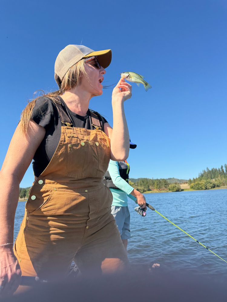 A woman in overalls holding up a small bass on a lake under a blue sky