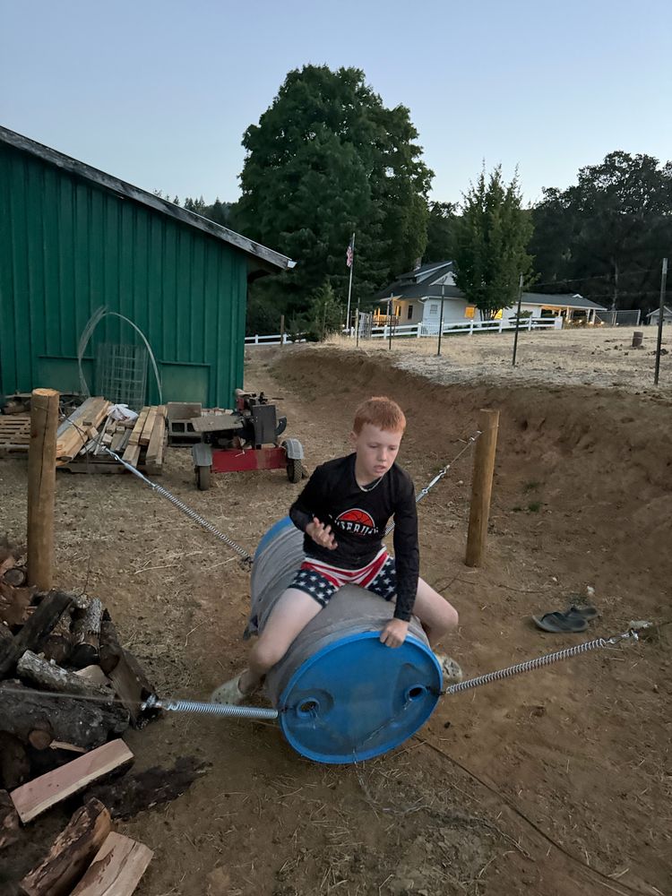 A boy riding a homemade bucking barrel on springs next to a green barn at dusk