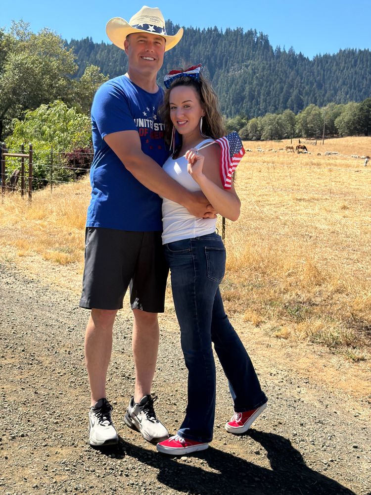 A man in a cowboy hat and a woman holding a small flag standing on a gravel road, with sheep and horses grazing in a dry pasture and a forested ridge behind